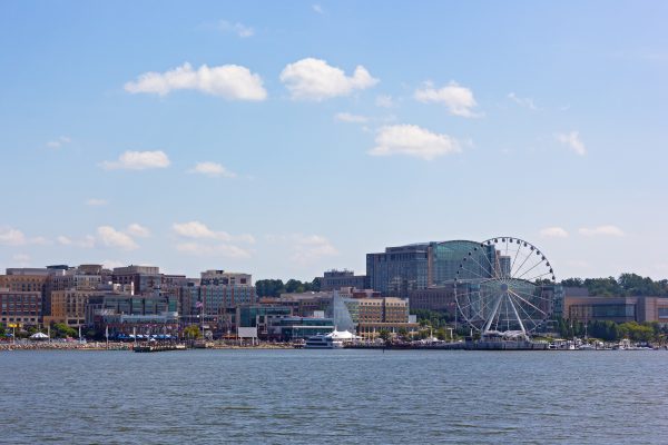 A waterfront view of a city skyline featuring modern buildings, a large Ferris wheel, and a bustling promenade along the water. The scene is set under a clear blue sky with a few clouds