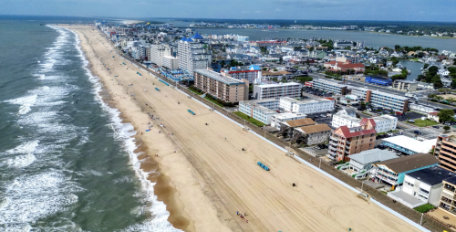 Aerial view of a beach with a wide sandy shoreline, bordered by a row of buildings and hotels. The ocean waves gently lap at the shore, and a boardwalk runs parallel to the beach. In the background, a coastal town with various structures and greenery is visible
