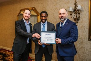 Three men are standing together in a formal setting, each wearing business attire. The man in the center is holding a certificate, while the other two men are positioned on either side, smiling and looking at the camera. The background features a decorative wall and a mirror