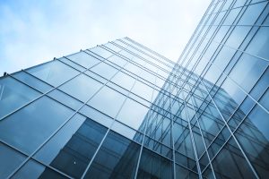 A modern glass building with a reflective facade, viewed from a low angle. The structure features sharp angles and a clear blue sky in the background