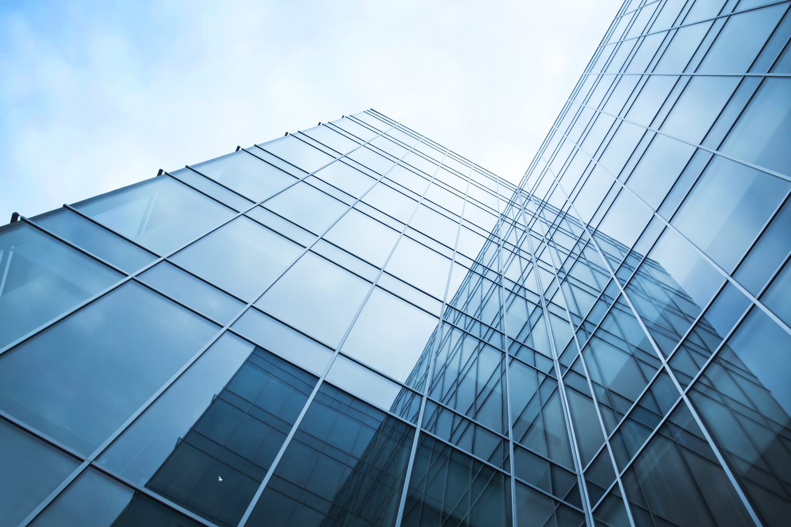 A modern glass building with a reflective facade, viewed from a low angle. The structure features sharp angles and a clear blue sky in the background