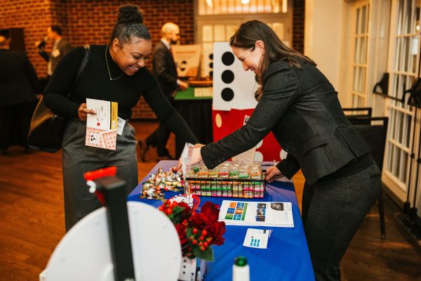Two women are interacting at a table filled with colorful gift boxes and promotional materials. One woman is reaching for an item while the other is holding a stack of papers and smiling. The setting appears to be a networking event or gathering, with people visible in the background