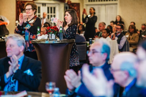A group of people are seated at tables in a banquet hall, with some clapping. In the foreground, several individuals are applauding, while a woman stands near a table holding a bouquet of red roses and looking towards the stage. Another woman is also standing and clapping. The setting appears to be a formal event or celebration
