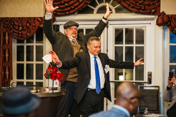 A man in a suit stands with his arms outstretched, while another man in a cap and vest stands behind him with his arms raised. The setting appears to be a formal event, with red curtains and a table decorated with flowers in the foreground. Several people are seated in the background, observing the scene