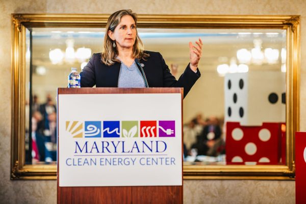 A woman stands at a podium with the Maryland Clean Energy Center logo, speaking to an audience. She has long brown hair and is wearing a gray top. A water bottle is placed on the podium, and there are decorative elements in the background, including a mirror and large dice