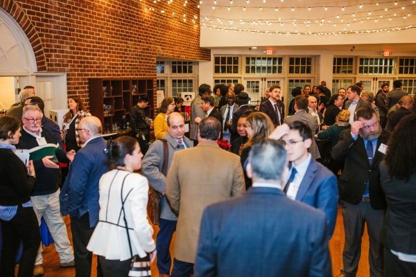 A crowded indoor event space with people engaged in conversation. The setting features brick walls and string lights overhead, creating a warm atmosphere. Attendees are dressed in business attire, with some holding drinks and others exchanging materials
