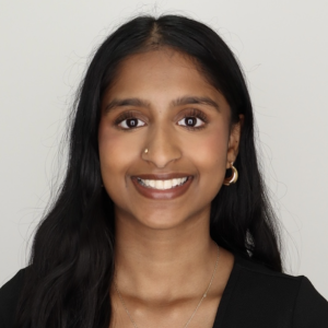 A young woman with long, straight black hair and a warm smile. She has a nose ring and is wearing hoop earrings. The background is plain and light-colored