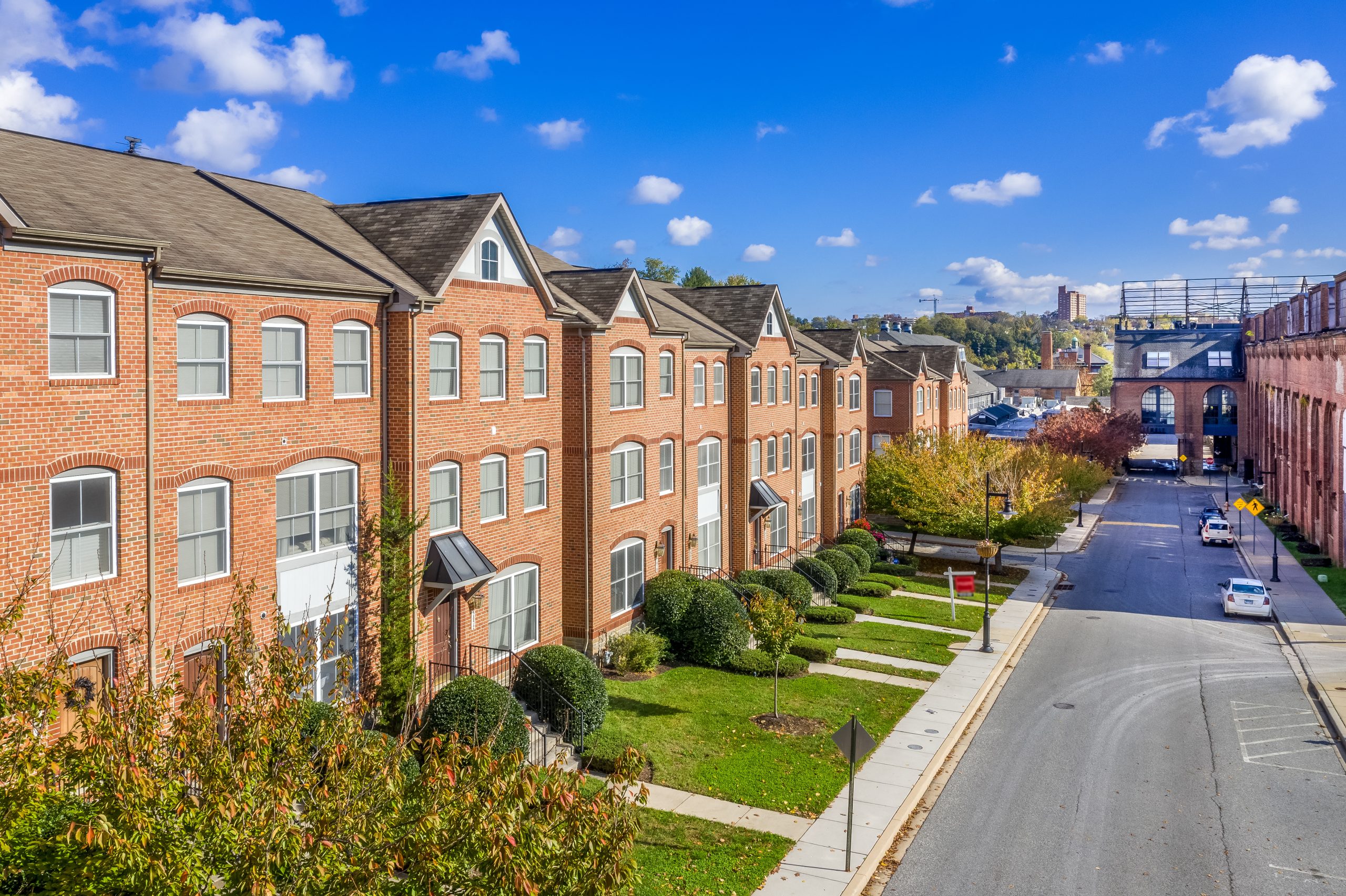 A row of brick townhouses with multiple windows and gabled roofs lines a well-maintained street. The scene features green lawns and neatly trimmed bushes in front of the houses. In the background, a bridge and trees are visible under a clear blue sky with scattered clouds