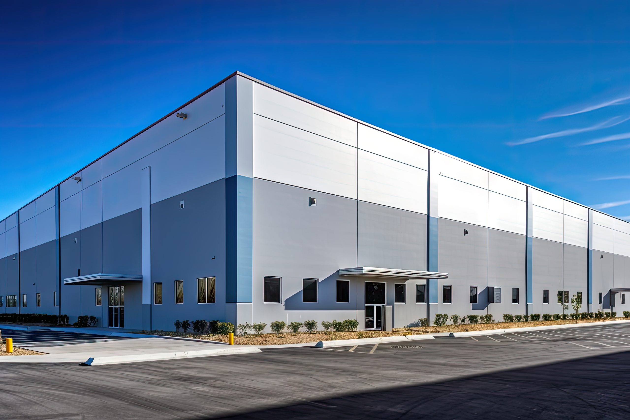 A large, modern industrial building with a gray and white exterior. The structure features a flat roof, multiple windows, and a covered entrance. The surrounding area includes a paved parking lot and landscaped greenery. The sky is clear and blue