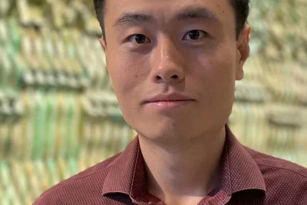 A young man with short black hair and a neutral expression stands in front of a textured, colorful background. He is wearing a maroon button-up shirt with a subtle pattern