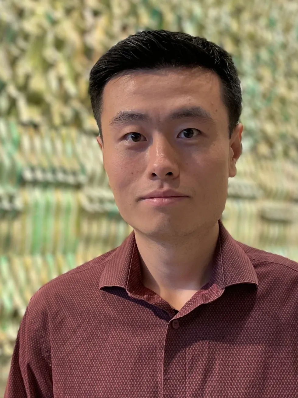 A young man with short black hair and a neutral expression stands in front of a textured, colorful background. He is wearing a maroon button-up shirt with a subtle pattern