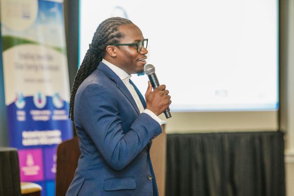 A man with long, braided hair speaks into a microphone while wearing a blue suit and glasses. He stands in front of a presentation screen, with a banner featuring various logos in the background