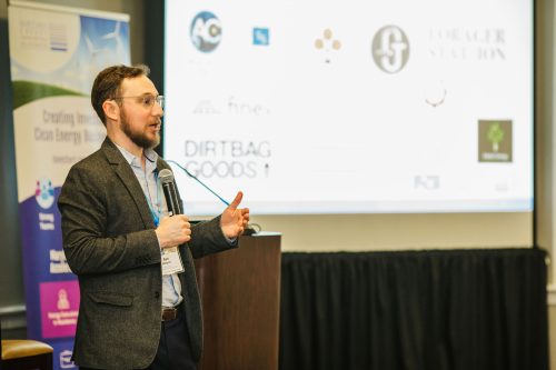 A man with glasses and a beard stands at a podium, speaking to an audience. He is wearing a gray blazer over a blue shirt. Behind him, a screen displays various logos and text related to a conference or event