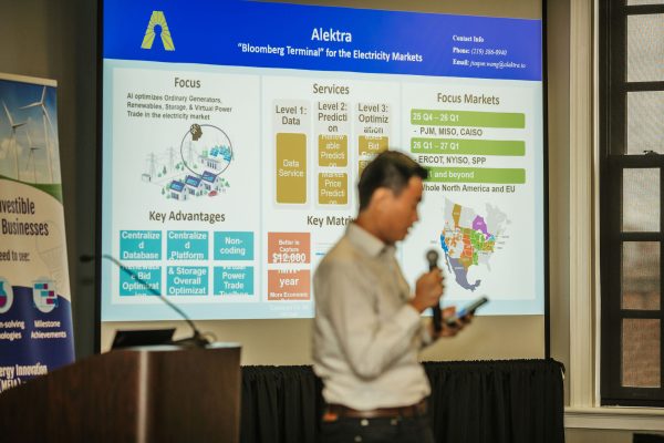 A person stands in the foreground holding a microphone, while a presentation screen in the background displays information about Alektra, including its focus, services, key advantages, and target markets. The screen features bullet points and a map highlighting regions in North America and the EU