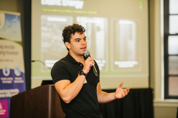 A young man with curly hair speaks into a microphone while gesturing with his hands. He is wearing a black shirt and stands in front of a presentation screen displaying information about energy. A podium is visible in the background
