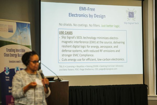 A woman stands in front of a presentation screen discussing EMI-free electronics designed to minimize electromagnetic interference. The screen displays the title "EMI-Free Electronics by Design" and outlines use cases in various industries, emphasizing energy efficiency and compliance