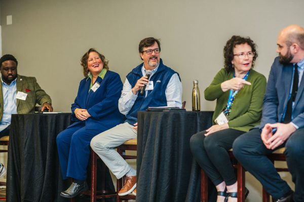 A panel discussion featuring four individuals seated at tables. One person is speaking into a microphone, while the others listen attentively. The panelists are dressed in business attire, and there are water bottles on the tables in front of them. The setting appears to be a conference or seminar