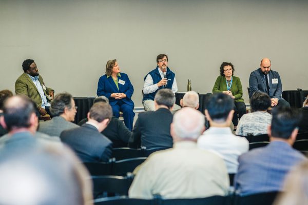 A panel discussion featuring five speakers seated at a table, with a diverse audience visible in the foreground. The speakers include three women and two men, each engaged in conversation. The setting appears to be a conference or seminar room