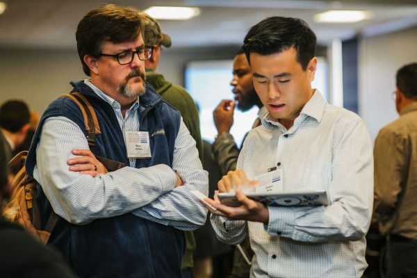 Two men are engaged in conversation at a networking event. One man, wearing glasses and a blue vest, appears to be listening intently, while the other man, dressed in a white shirt, is taking notes on a tablet. In the background, other attendees are visible, suggesting a busy atmosphere
