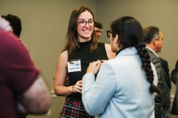A woman with long brown hair and glasses smiles while holding a drink. She is wearing a black sleeveless top and a plaid skirt. Another woman with dark hair in a braid is talking to her. The setting appears to be a professional networking event