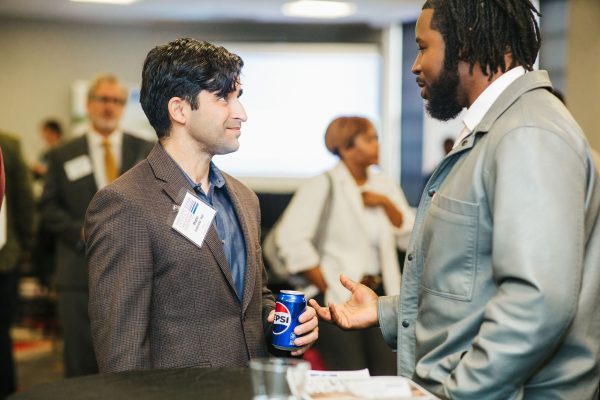 Two men are engaged in conversation at a networking event. One man, wearing a brown blazer, holds a can of Pepsi and smiles while speaking. The other man, dressed in a light gray jacket, listens attentively. In the background, other attendees are visible, some mingling and others in conversation