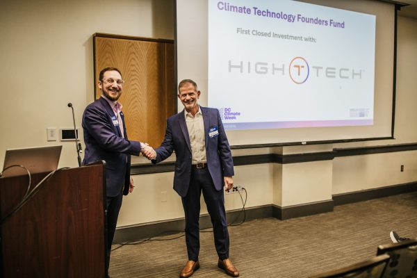 Two men in business attire are shaking hands in a conference room. One man is standing at a podium, while the other stands beside him. A presentation screen in the background displays the text "Climate Technology Founders Fund" and "First Closed Investment with: HIGHTECH