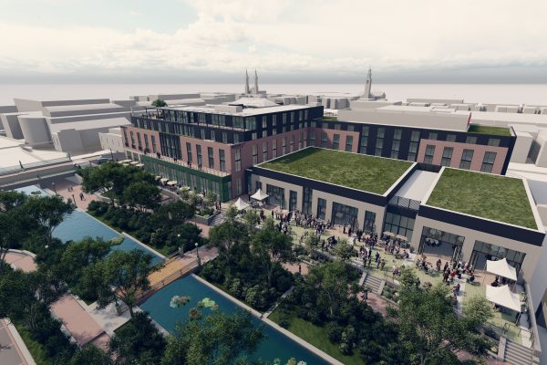 A modern multi-story building with a green roof is situated near a landscaped area featuring trees and a pond. People are gathered outside, likely for an event, with tables and tents set up. The surrounding area includes additional buildings and a clear sky above