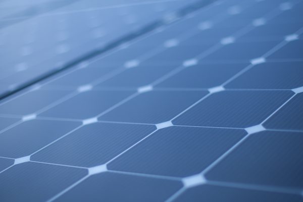 Close-up view of solar panels, showcasing a grid of dark photovoltaic cells with a textured surface and a geometric pattern. The panels are arranged in rows, reflecting light