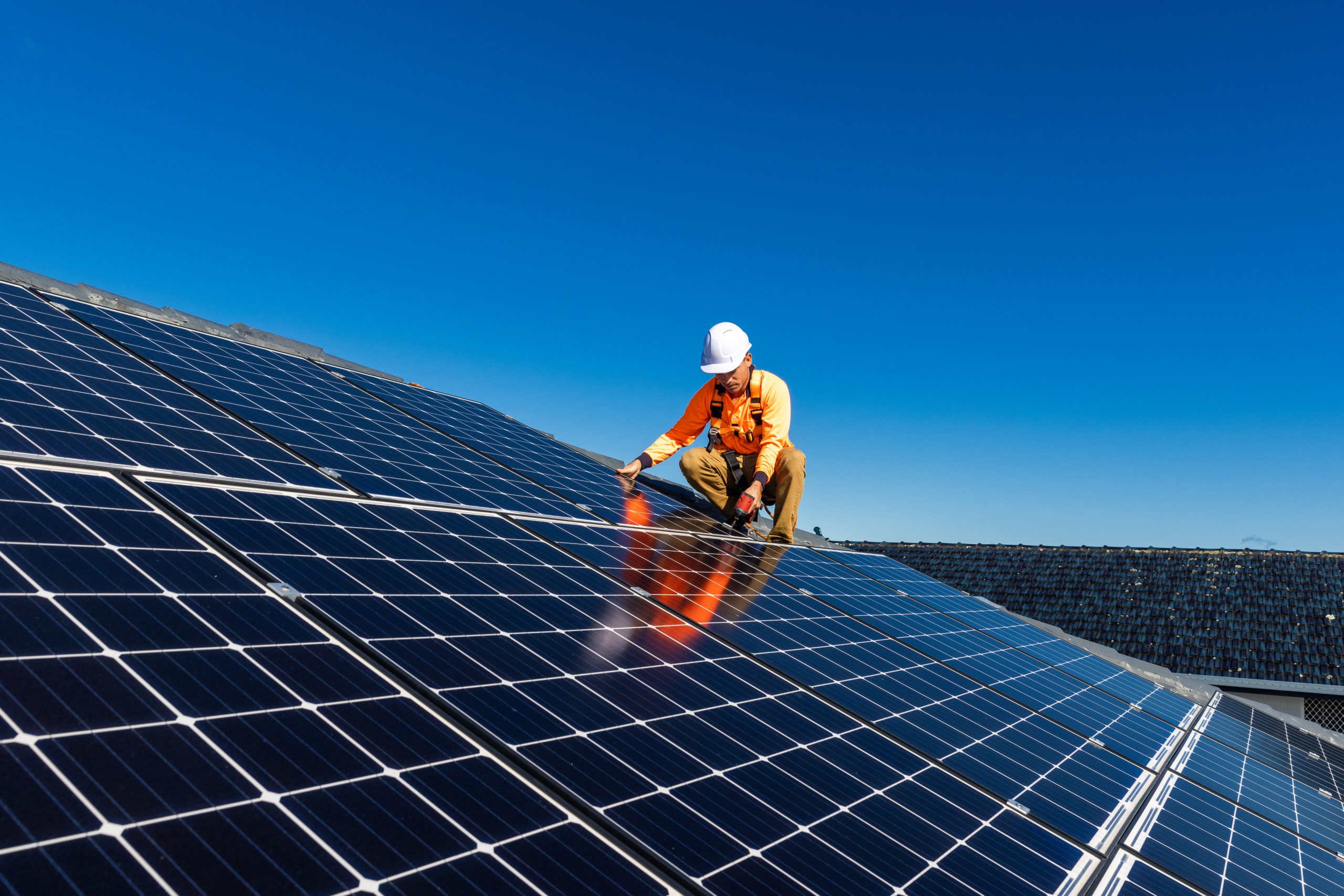 A worker in an orange shirt and white hard hat is installing solar panels on a rooftop under a clear blue sky