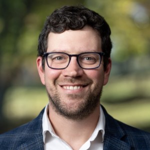 A man with curly dark hair and glasses smiles at the camera. He is wearing a blue blazer over a white shirt, and the background features blurred greenery