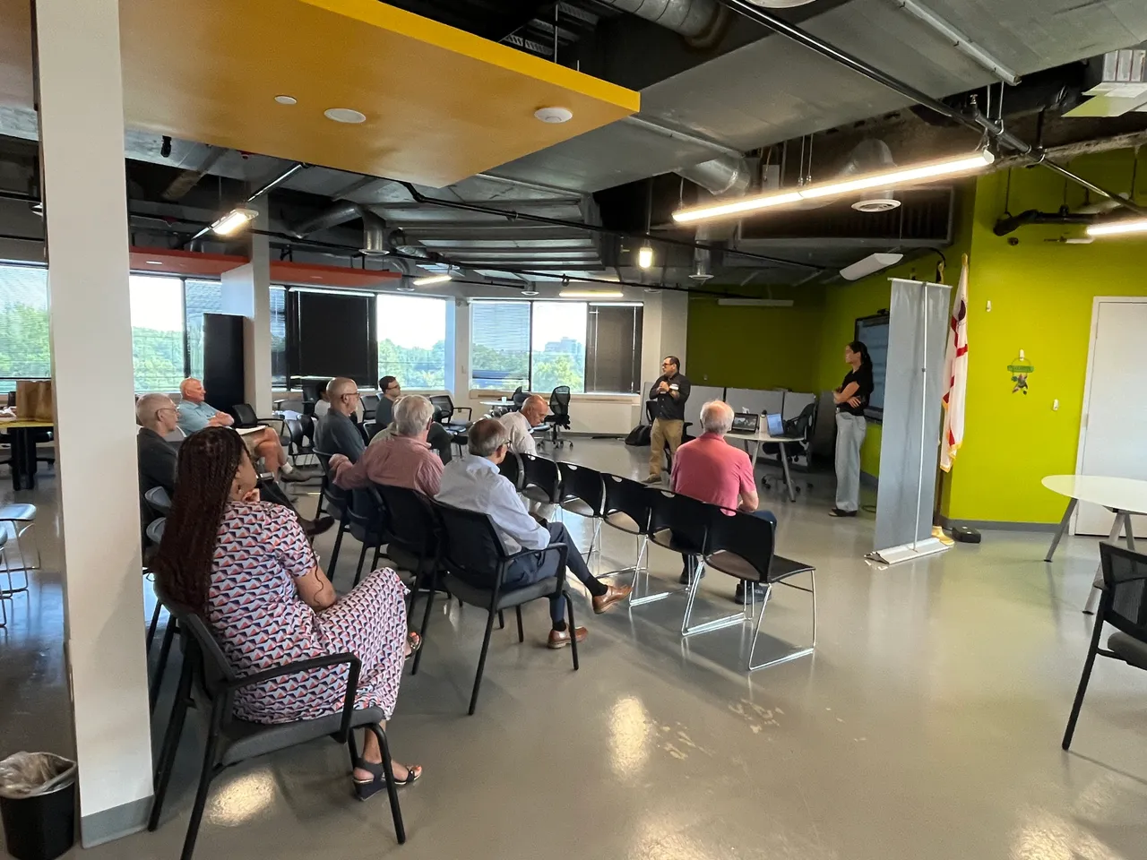A group of people sits in chairs facing two speakers in a modern indoor space. The audience, consisting of mostly older adults, is engaged in a discussion. The room features large windows, bright green walls, and a mix of seating arrangements