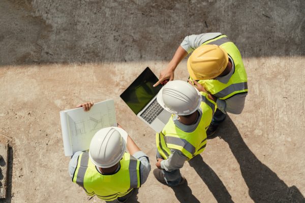 Three construction workers wearing yellow hard hats and reflective vests are gathered around a laptop on a concrete surface. One worker is pointing at the laptop screen while another holds a set of blueprints