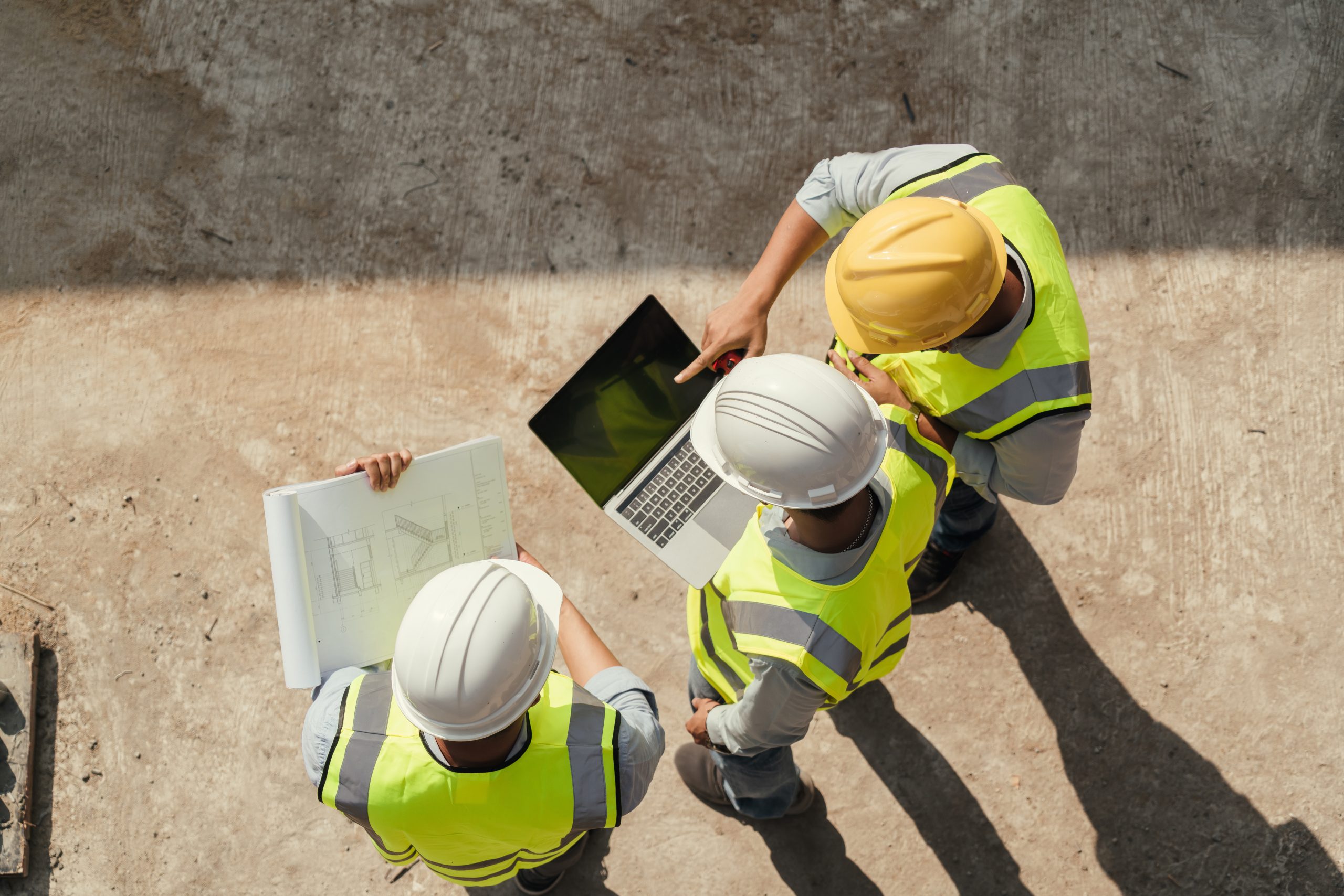 Three construction workers wearing yellow hard hats and reflective vests are gathered around a laptop on a concrete surface. One worker is pointing at the laptop screen while another holds a set of blueprints