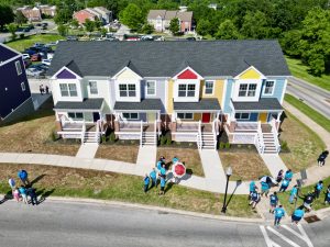 A row of four colorful townhouses with porches, each painted in different pastel shades. In front of the houses, a group of people wearing blue shirts gathers on the grass, with some holding umbrellas. The scene is set in a suburban area with additional buildings and trees in the background