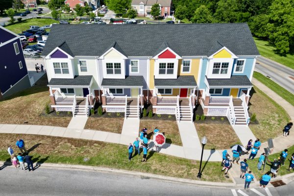 A row of four colorful townhouses with porches, each painted in different pastel shades. In front of the houses, a group of people wearing blue shirts gathers on the grass, with some holding umbrellas. The scene is set in a suburban area with additional buildings and trees in the background