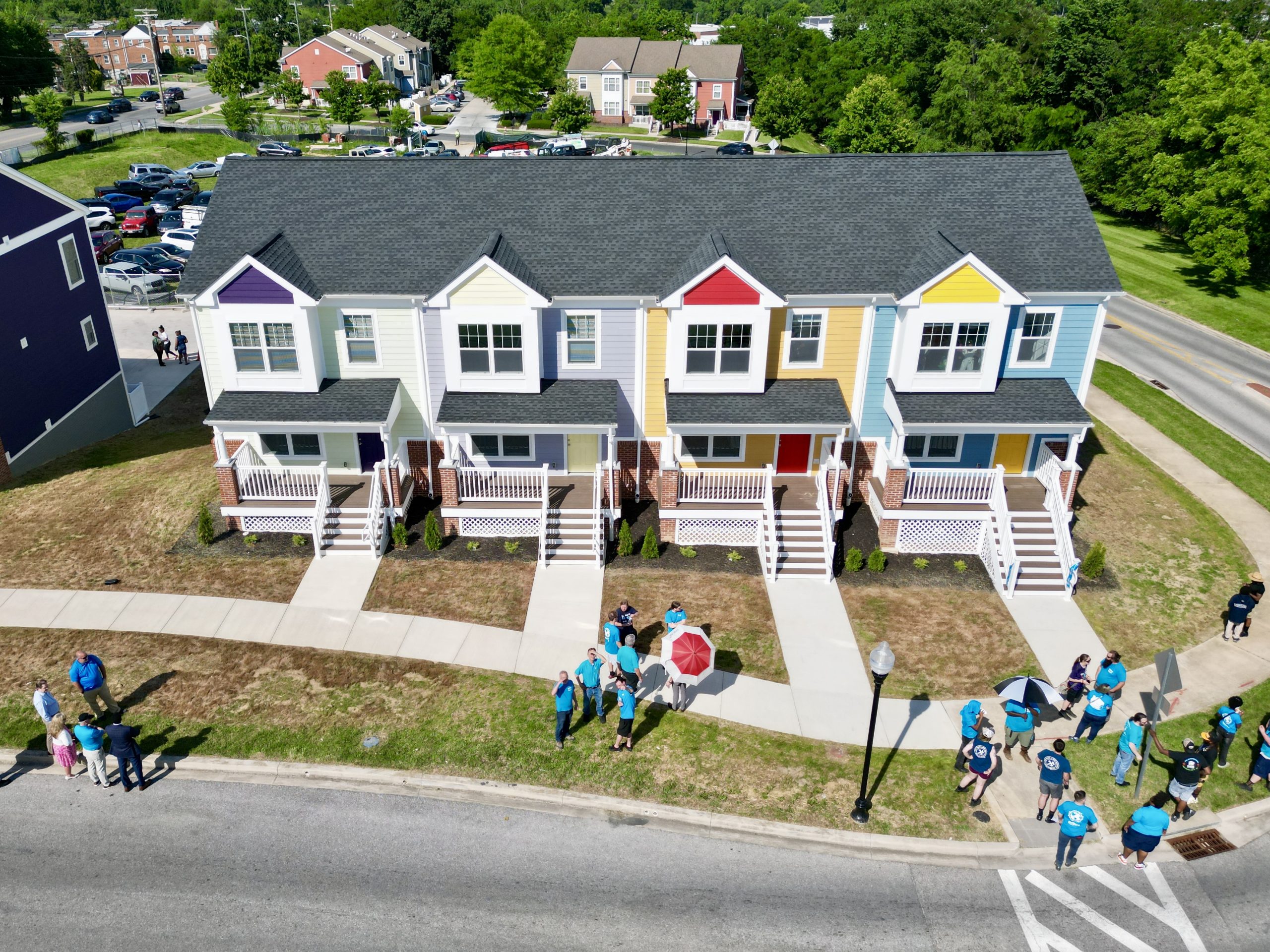 A row of four colorful townhouses with porches, each painted in different pastel shades. In front of the houses, a group of people wearing blue shirts gathers on the grass, with some holding umbrellas. The scene is set in a suburban area with additional buildings and trees in the background