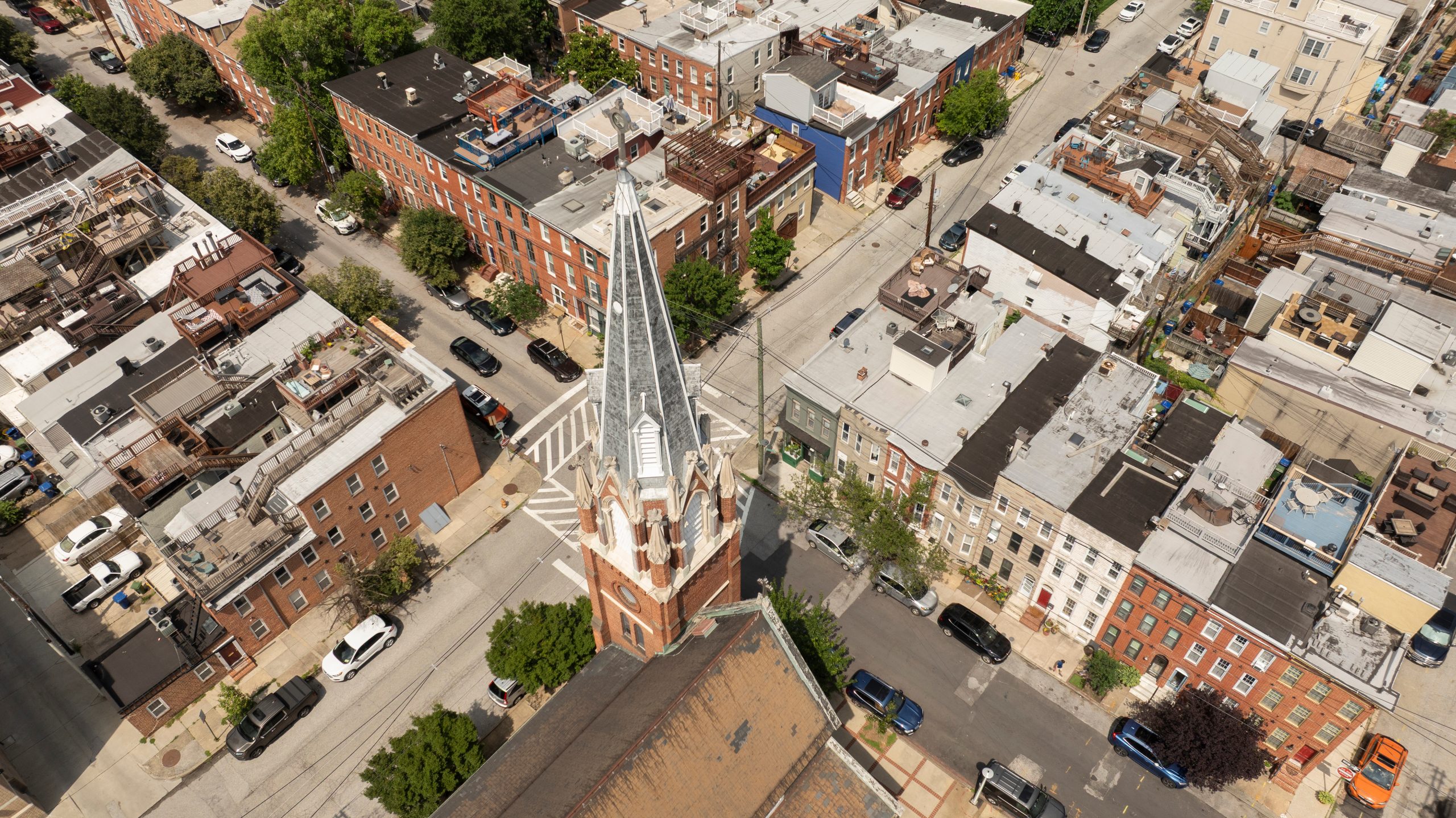 Aerial view of a city block featuring a prominent church with a tall steeple at the center. Surrounding the church are residential buildings with flat roofs and a mix of brick and painted facades. Streets are visible, lined with parked cars and greenery