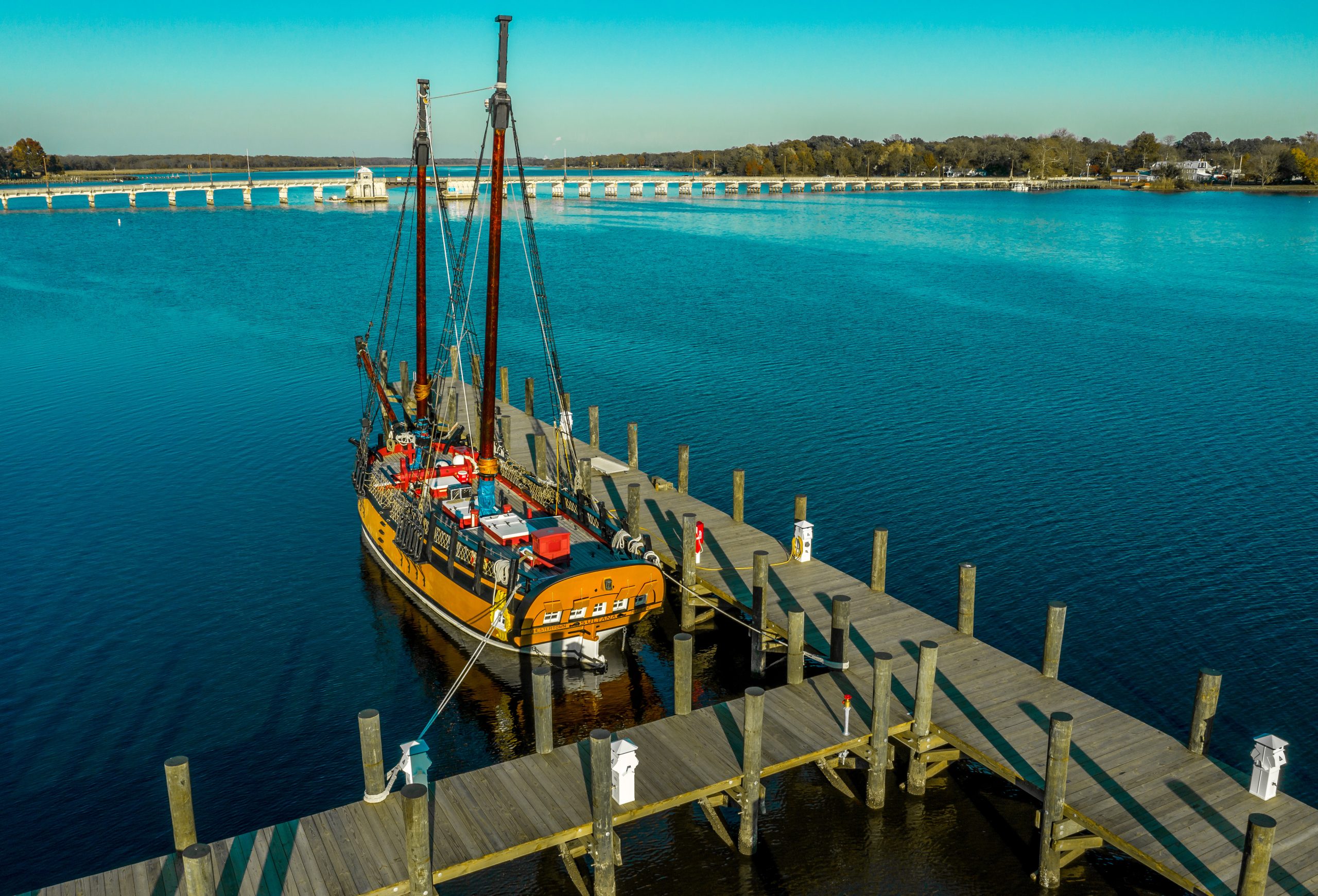 A wooden sailing ship with multiple masts is docked at a wooden pier. The water is calm and reflects the clear blue sky. In the background, a bridge spans the waterway, and trees line the shore