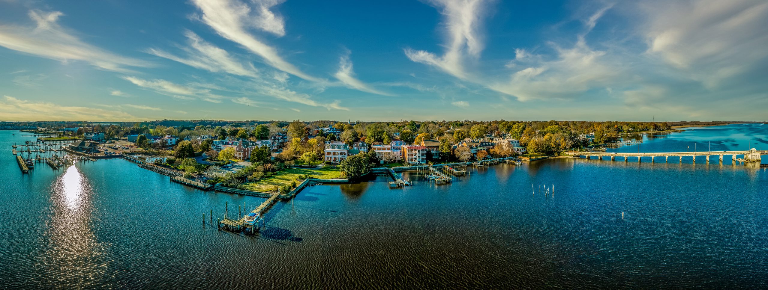 A panoramic view of a coastal town featuring a mix of residential buildings and docks along the water. The scene includes boats in the water, trees with autumn foliage, and a clear blue sky with scattered clouds. The shoreline is lined with piers and grassy areas