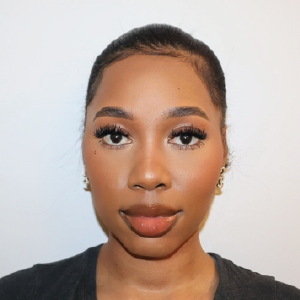 A woman with long, dark hair styled in a bun is facing the camera. She has a smooth complexion, wearing makeup that includes long eyelashes and a nude lip color. She is dressed in a dark shirt and has small, decorative earrings. The background is plain and white