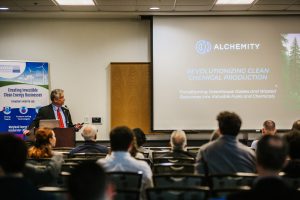 A speaker stands at a podium in front of an audience, presenting a slide titled "Revolutionizing Clean Chemical Production" by Alchemy. The slide highlights the transformation of greenhouse gases and waste resources into valuable fuels and chemicals. The setting appears to be a conference or seminar focused on clean energy and environmental innovation