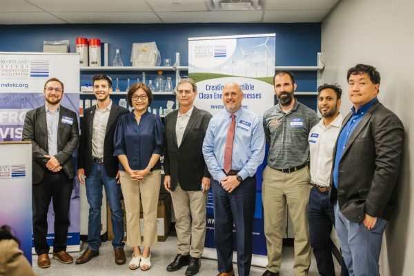 A group of seven individuals stands together in a laboratory setting, smiling at the camera. They are dressed in business casual attire and are positioned in front of banners promoting clean energy initiatives. The background features laboratory equipment and shelves with various scientific materials