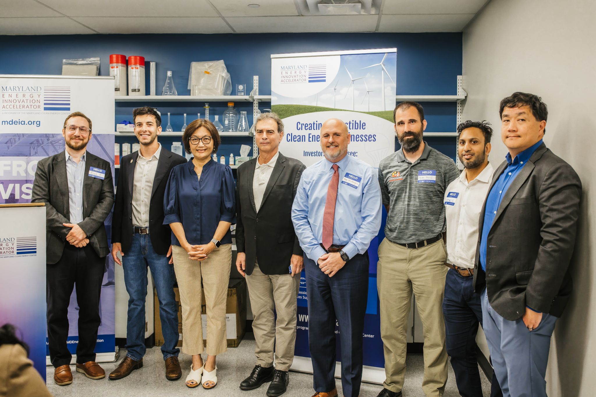 A group of seven individuals stands together in a laboratory setting, smiling at the camera. They are dressed in business casual attire and are positioned in front of banners promoting clean energy initiatives. The background features laboratory equipment and shelves with various scientific materials