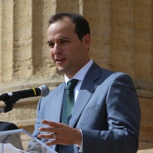 A man in a gray suit and green tie stands at a podium with a microphone, speaking. He gestures with his right hand and appears to be addressing an audience. The background features stone columns