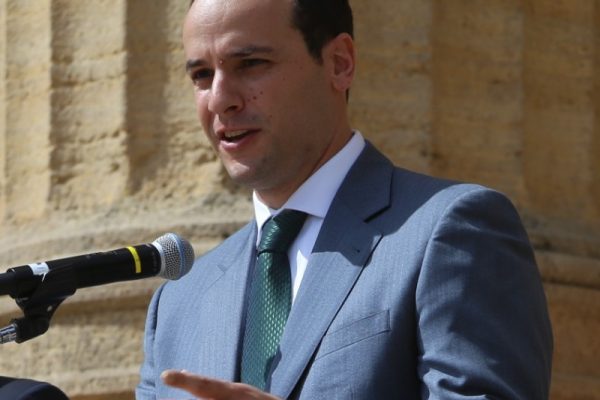 A man in a gray suit and green tie stands at a podium with a microphone, speaking. He gestures with his right hand and appears to be addressing an audience. The background features stone columns