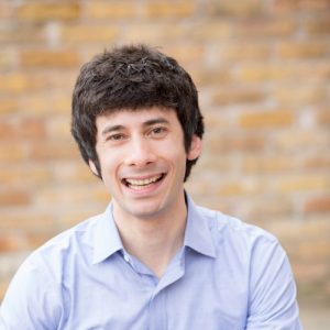 A young man with dark, curly hair smiles at the camera, wearing a light blue button-up shirt. He is seated in front of a brick wall with a warm, neutral tone
