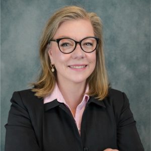 A woman with shoulder-length blonde hair and glasses smiles while wearing a black blazer over a light pink shirt. The background is a textured gray