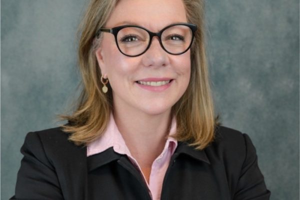 A woman with shoulder-length blonde hair and glasses smiles while wearing a black blazer over a light pink shirt. The background is a textured gray