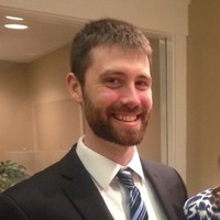 A man with a beard and short hair smiles while wearing a suit and tie. He is indoors, with soft lighting and a neutral background
