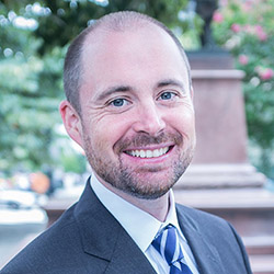 A man with short, light brown hair and a beard smiles while wearing a dark suit and a striped tie. He is standing outdoors with trees and a monument in the background